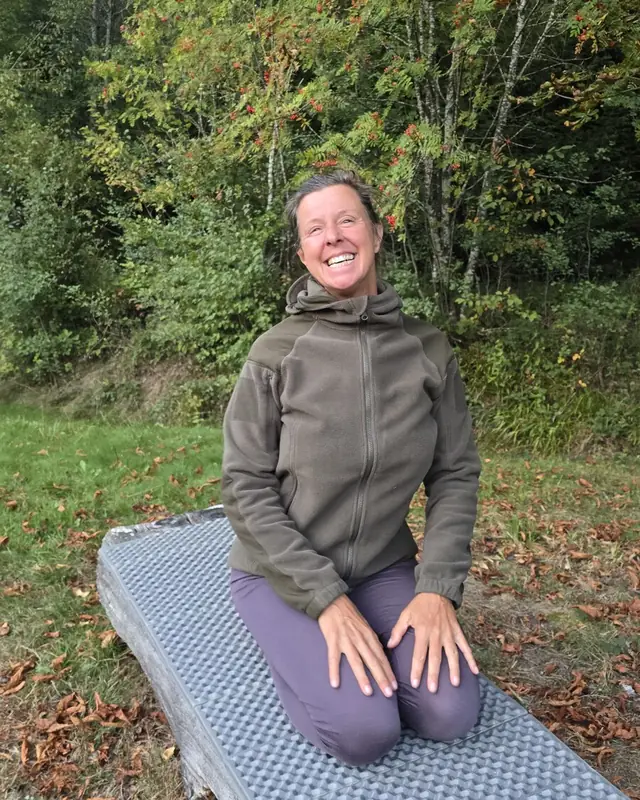 Christina kniet entspannt auf einer Matte auf einem Baumstamm und lächelt