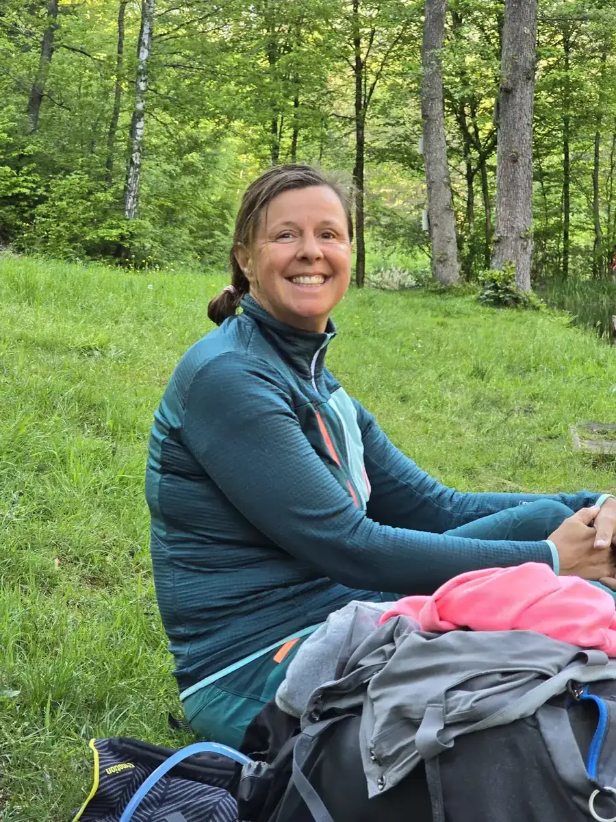Christina sitzt lächelnd mit Rucksack auf einer Wiese am Waldrand