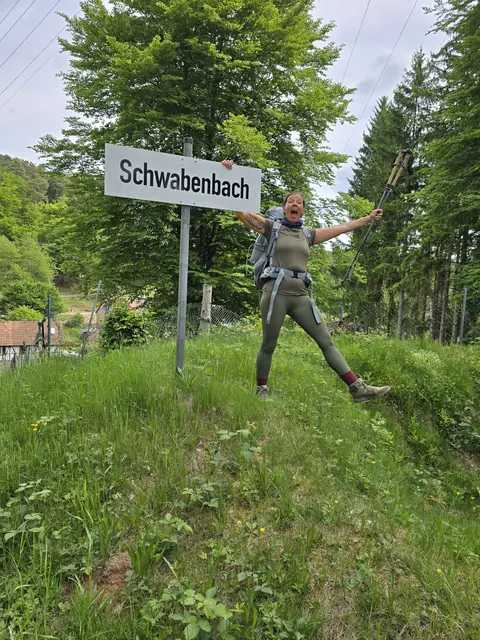 Christina jubelt mit Wanderstöcken neben dem Ortsschild Schwabenbach