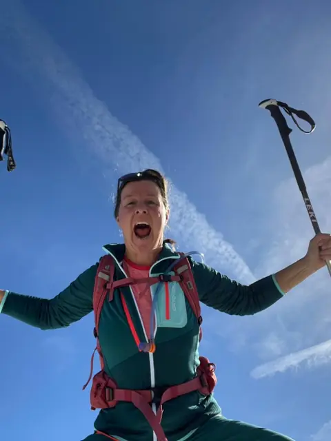 Christina lacht von unten fotografiert in den blauen Himmel und reckt die Stöcke hoch